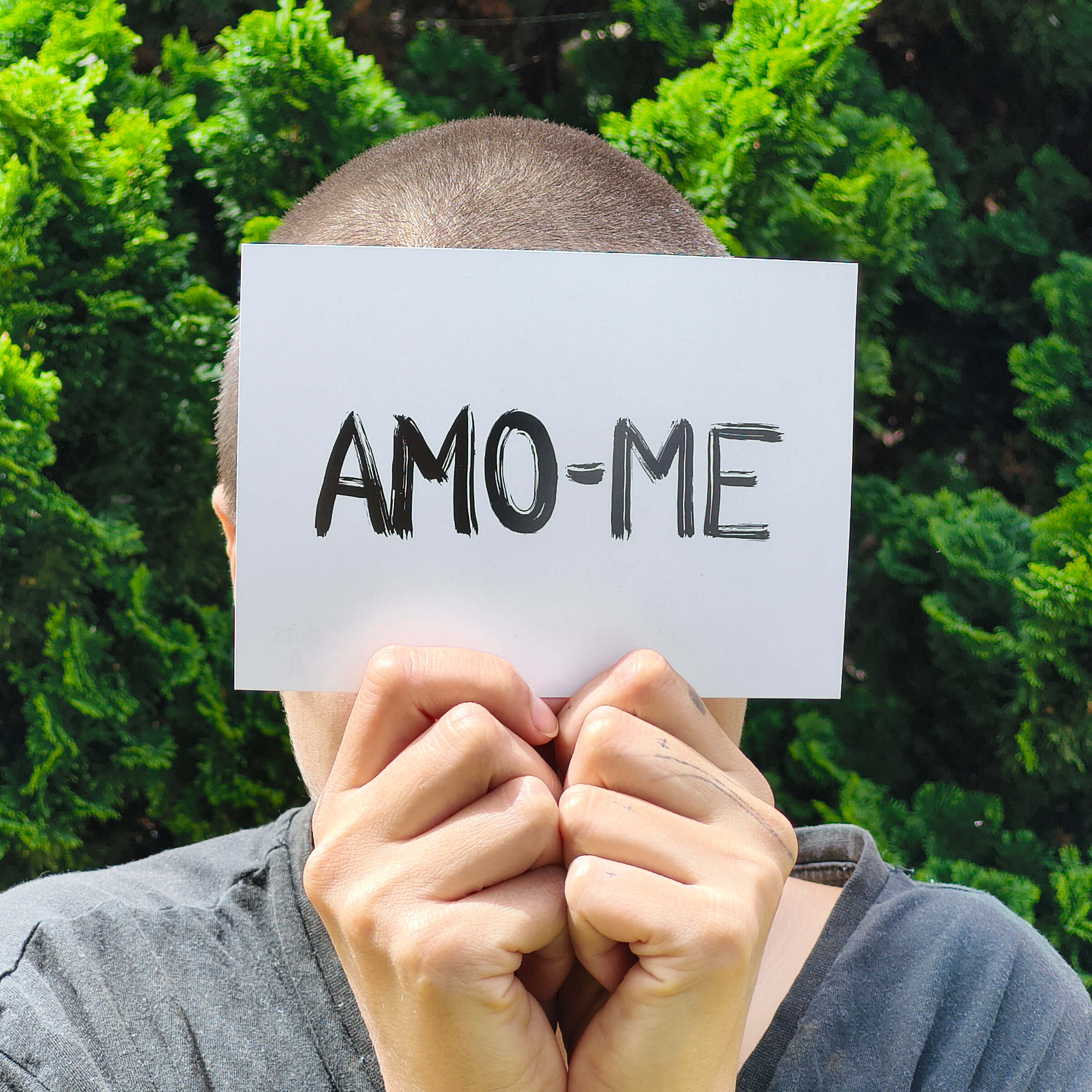 person with very short hair holding a postcard with amo-me handwritten on it, which means I love myself in Portuguese